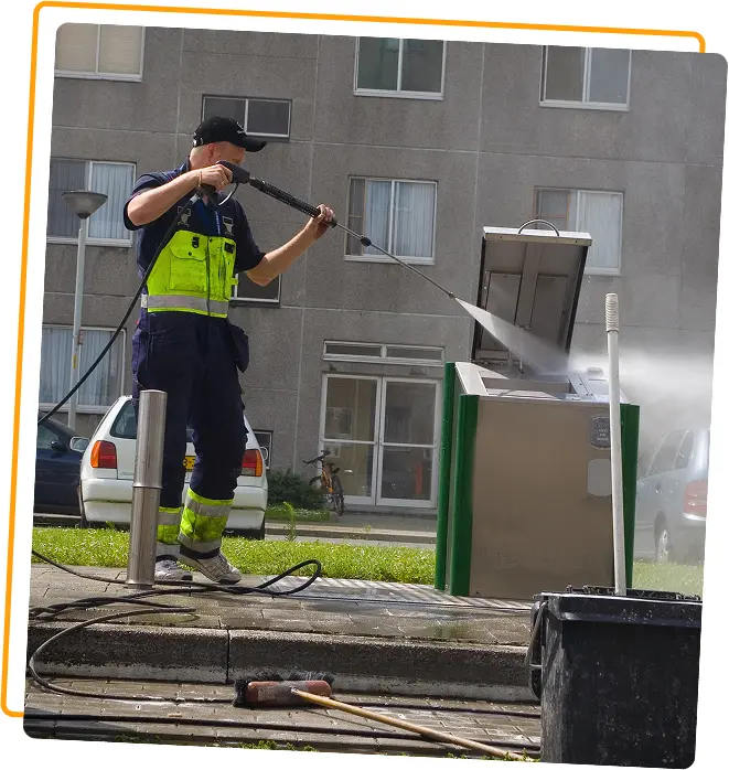 Worker pressure washing trash container outdoors.
