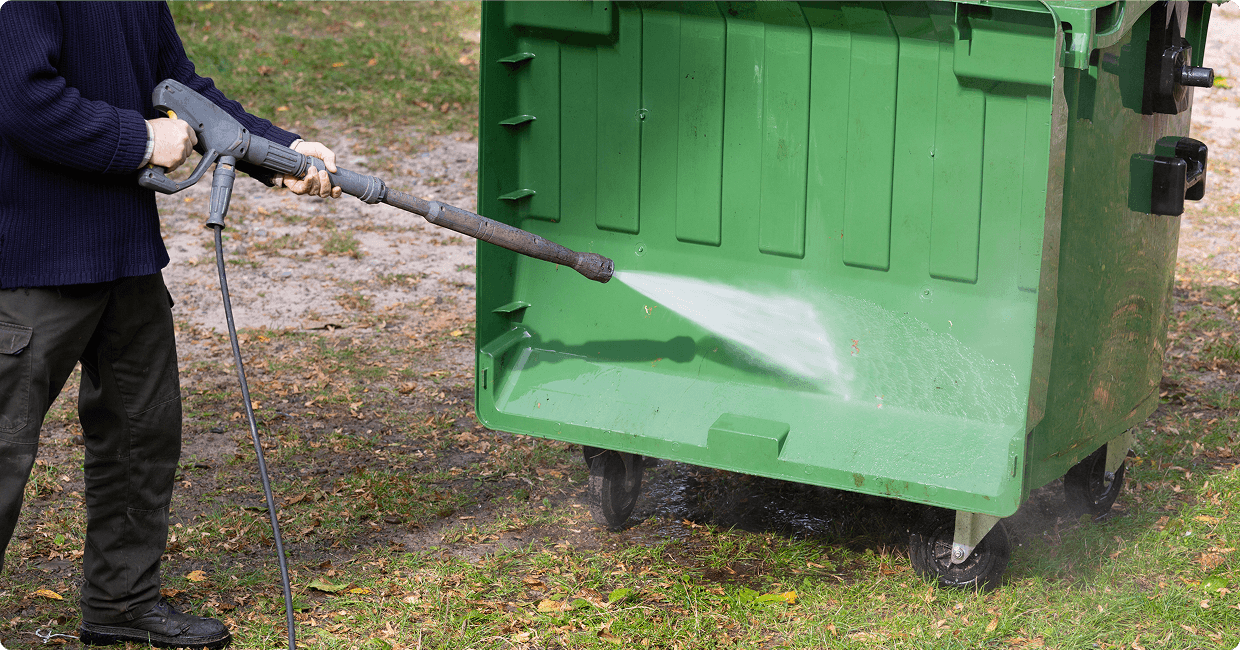 Person pressure washing a green dumpster.